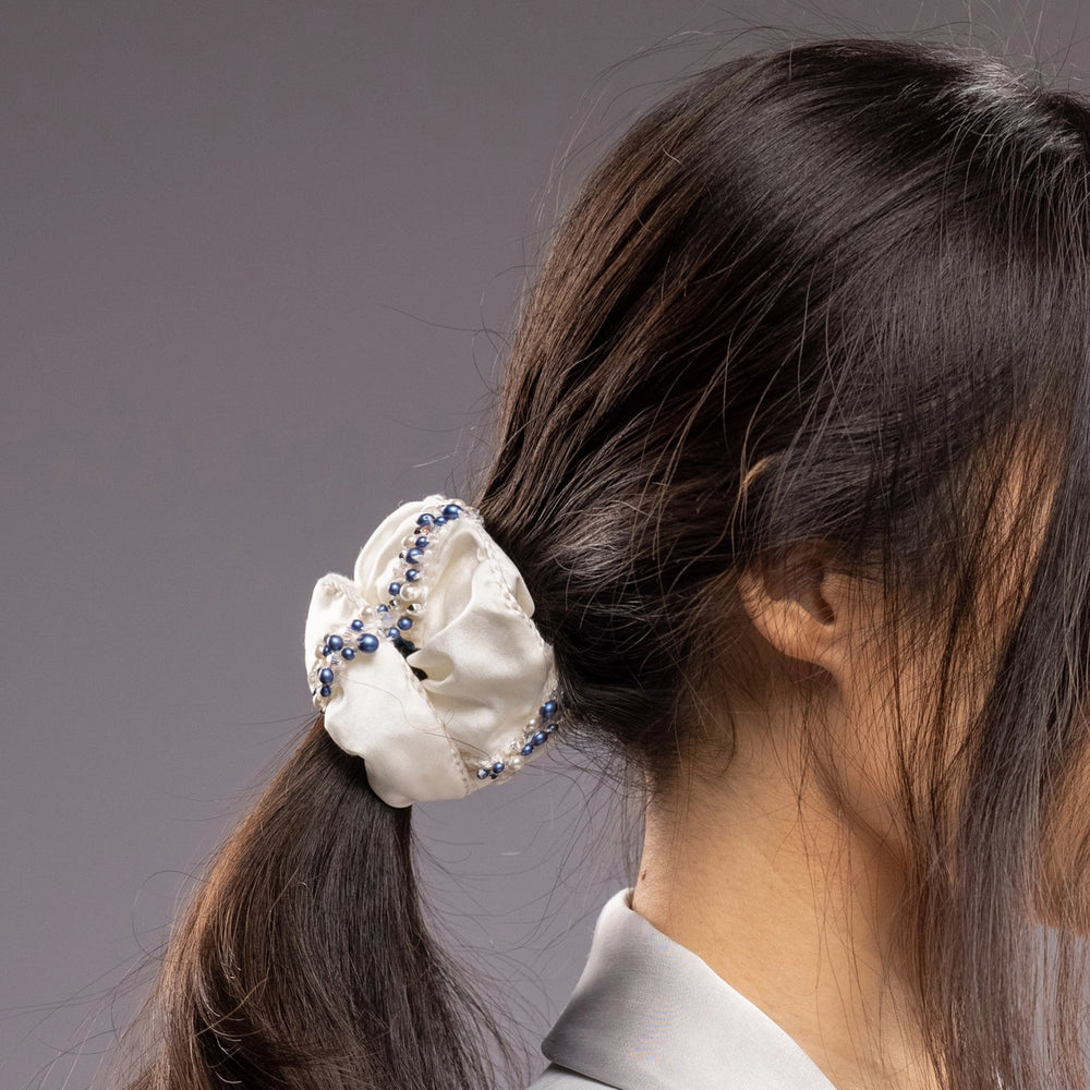 
                  
                    A dark-haired woman in grey jacket, view from the side, with a stylish ponytail is wearing Malkiele designer white silk scrunchie, embellished with blue pearls, details, name Elizabeth
                  
                