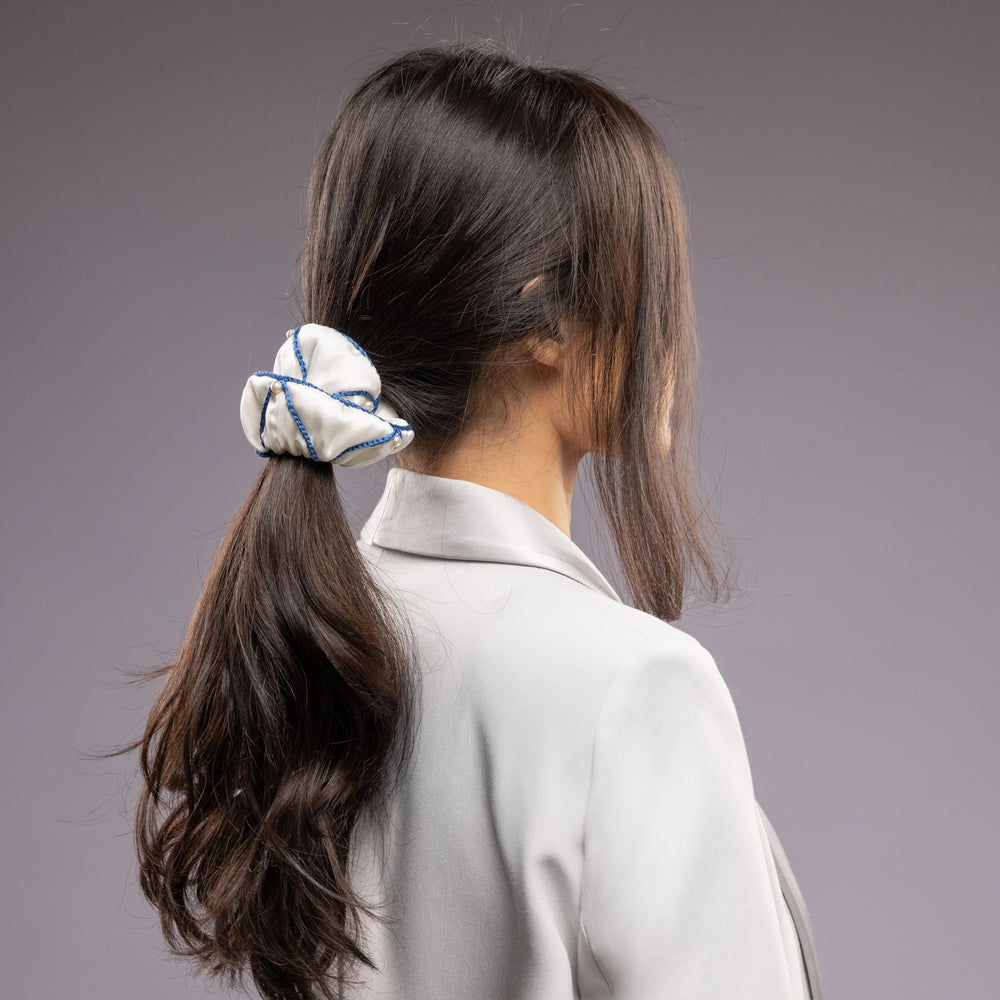 
                  
                    A black-haired woman in grey jacket, view from the back, with a stylish ponytail is wearing Malkiele designer white silk scrunchie, embellished with white pearls and blue silk knit ribbon, name Majestic.
                  
                