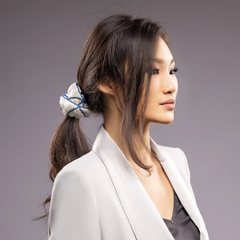 
                  
                    A black-haired woman in grey jacket, front view, with a stylish ponytail is wearing Malkiele designer white silk scrunchie, embellished with white pearls and blue silk knit ribbon, name Majestic.
                  
                