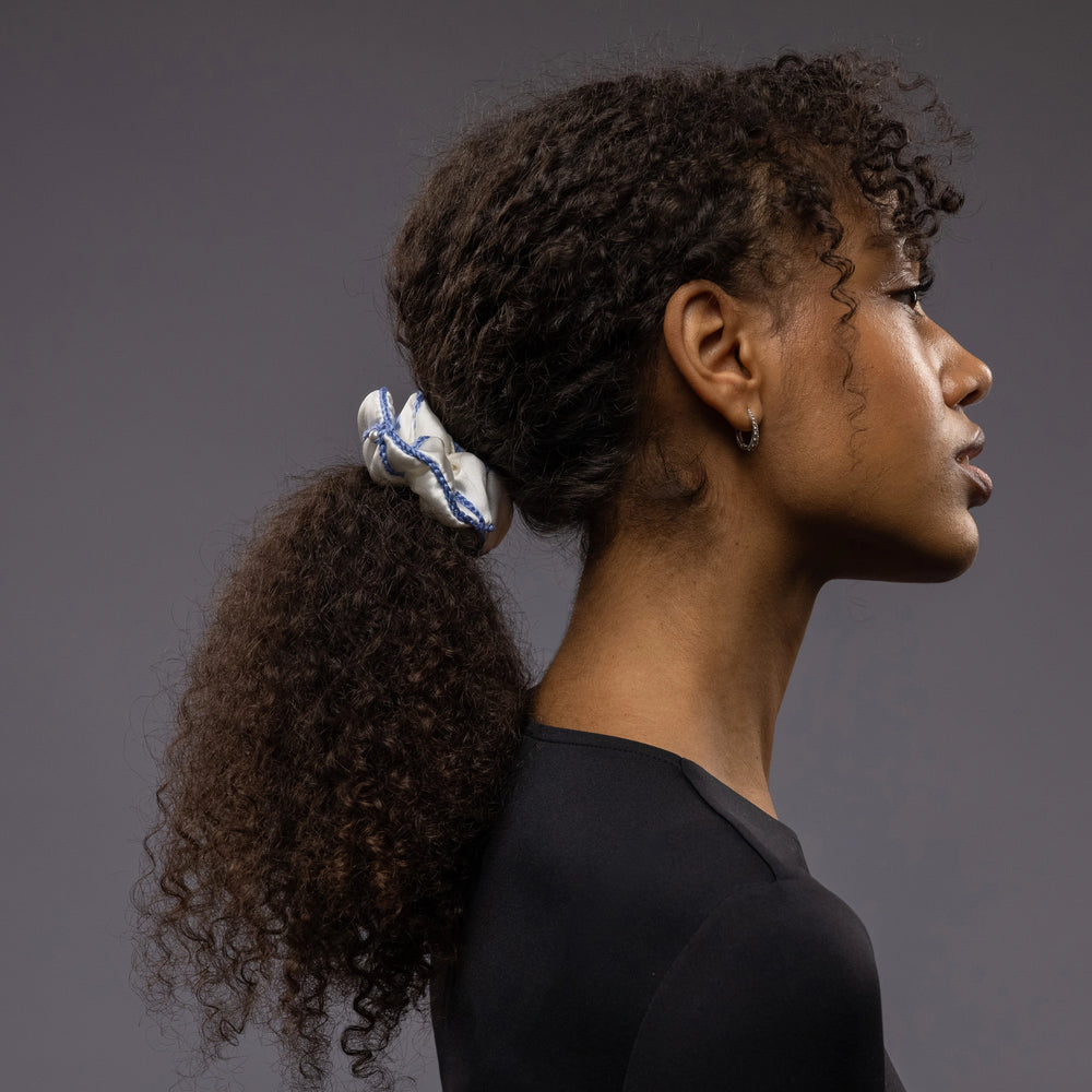 
                  
                    African woman, the view from the side, with a stylish ponytail is wearing Malkiele designer white silk scrunchie, embellished with white pearls and light blue silk knit ribbon, name Majestic.
                  
                