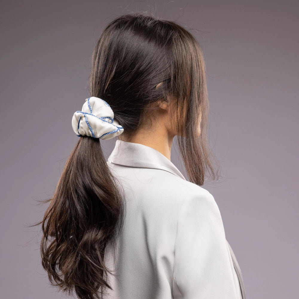 
                  
                    A black-haired woman in grey jacket, the view from the back, with a stylish ponytail is wearing Malkiele designer white silk scrunchie, embellished with white pearls and light blue silk knit ribbon, name Majestic.
                  
                