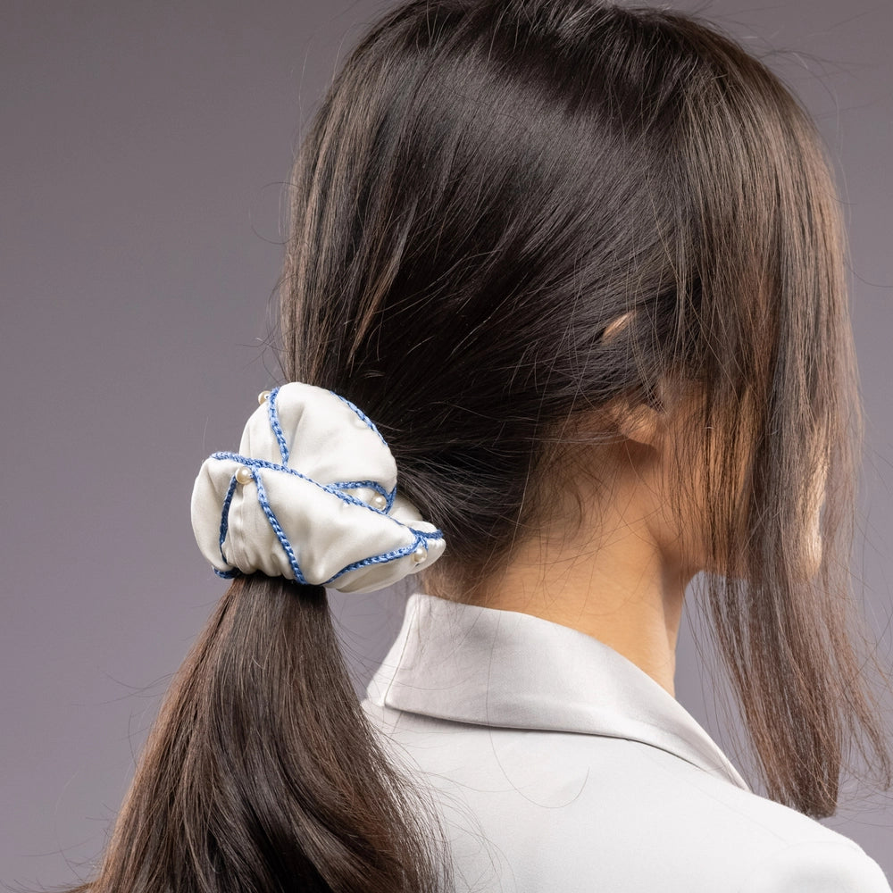 
                  
                    A black-haired woman, the view from the back, with a stylish ponytail is wearing Malkiele designer white silk scrunchie, embellished with white pearls and light blue silk knit ribbon, details, name Majestic.
                  
                