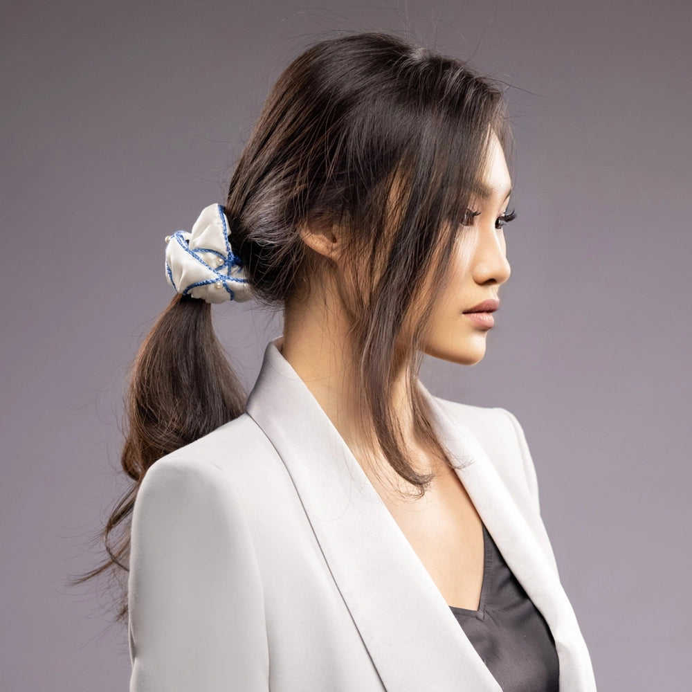 
                  
                    Asian woman in a grey jacket, front view, with a stylish ponytail is wearing Malkiele designer white silk scrunchie, embellished with white pearls and light blue silk knit ribbon, name Majestic.
                  
                