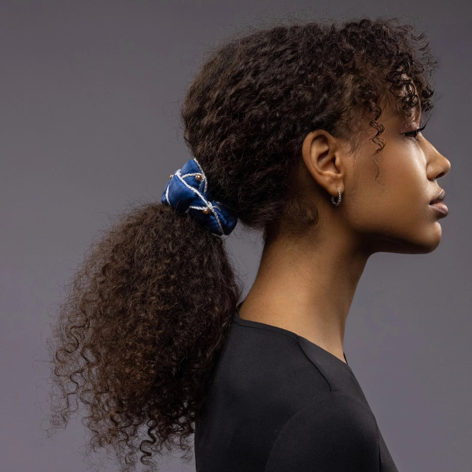 
                  
                    African woman, side view, with a stylish ponytail is wearing Malkiele designer royal blue silk scrunchie, embellished with gold pearls and white silk knit ribbon, name Majestic.
                  
                