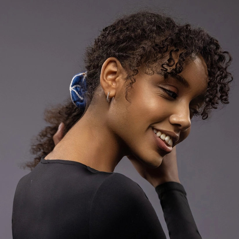 
                  
                    Smiling African woman, front view, with a stylish ponytail is wearing Malkiele designer royal blue silk scrunchie, embellished with gold pearls and white silk knit ribbon, name Majestic.
                  
                