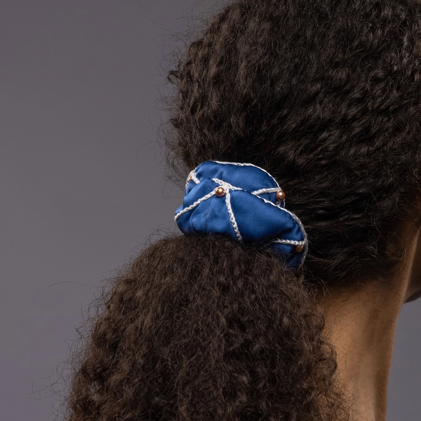 
                  
                    African woman, back view, details, with a stylish ponytail is wearing Malkiele designer royal blue silk scrunchie, embellished with gold pearls and white silk knit ribbon, name Majestic.
                  
                