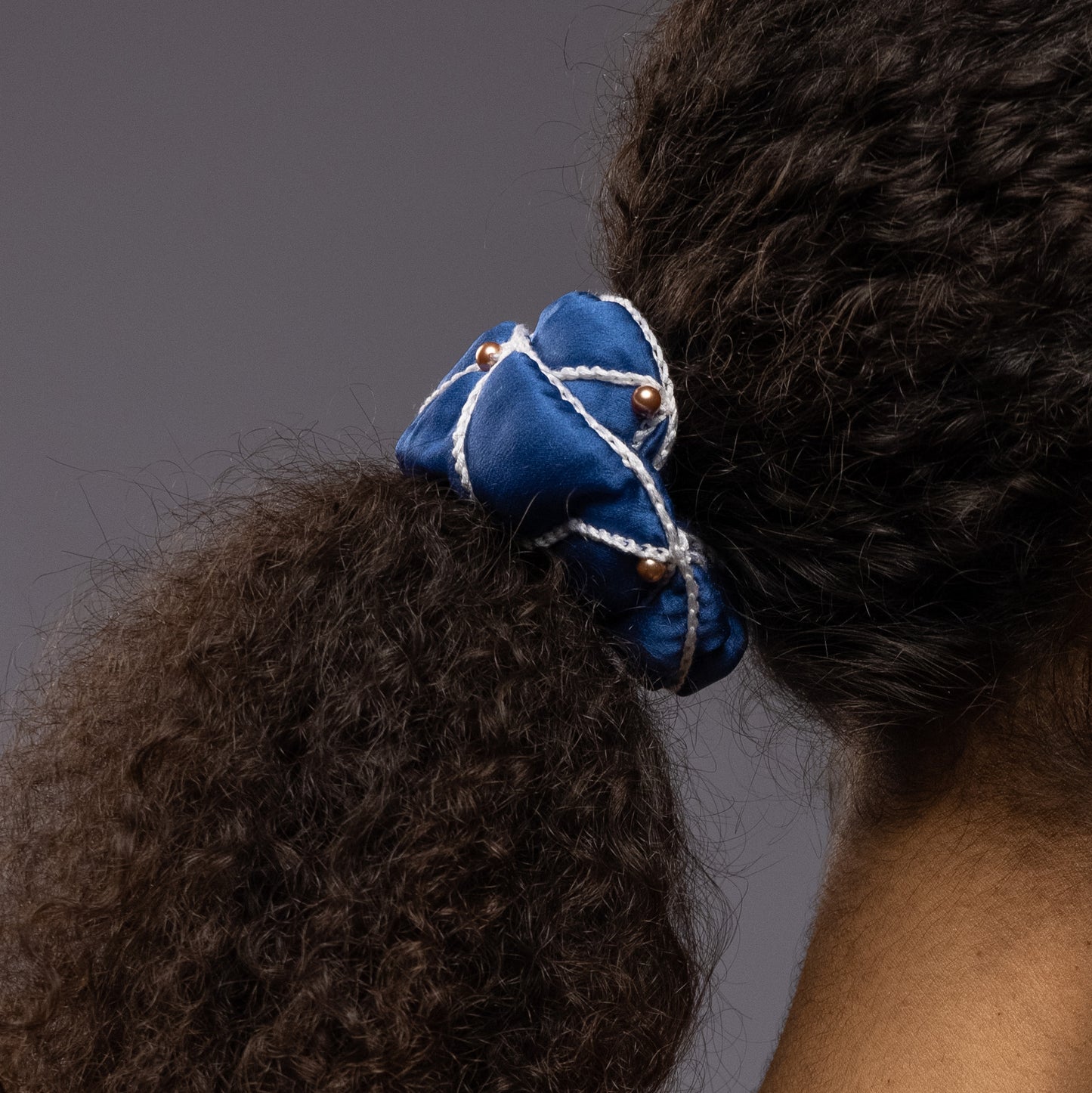 
                  
                    African woman, side view, details, with a stylish ponytail is wearing Malkiele designer royal blue silk scrunchie, embellished with gold pearls and white silk knit ribbon, name Majestic.
                  
                