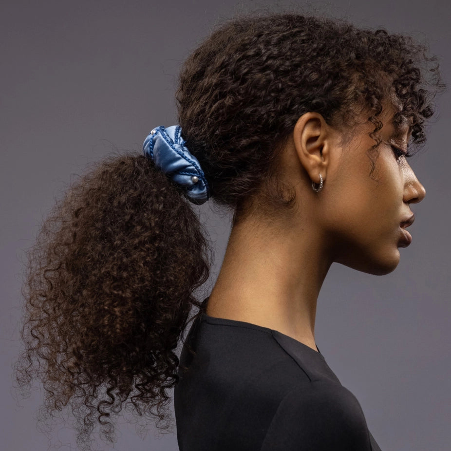 A dark-haired girl, side view, with stylish ponytail is wearing light blue Malkiele designer silk scrunchie, embellished with pearls, name Majestic.