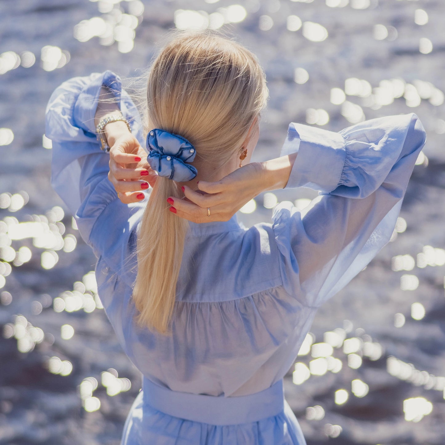 A blonde-haired girl in light blue dress, back view, is touching with hands her ponytail with light blue Malkiele designer silk scrunchie, name Majestic.