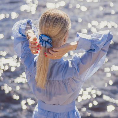 A blonde-haired girl in light blue dress, back view, is touching with hands her ponytail with light blue Malkiele designer silk scrunchie, name Majestic.