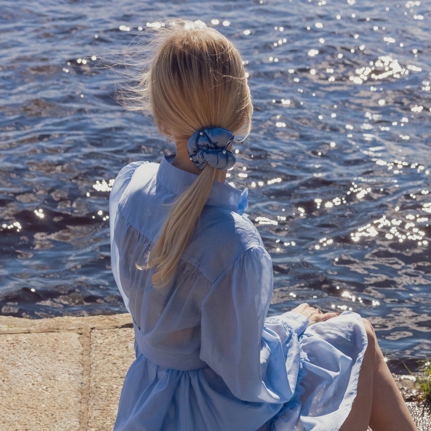 
                  
                    A blonde-haired girl in light blue dress, back view, with stylish ponytail is wearing light blue Malkiele designer silk scrunchie, name Majestic.
                  
                