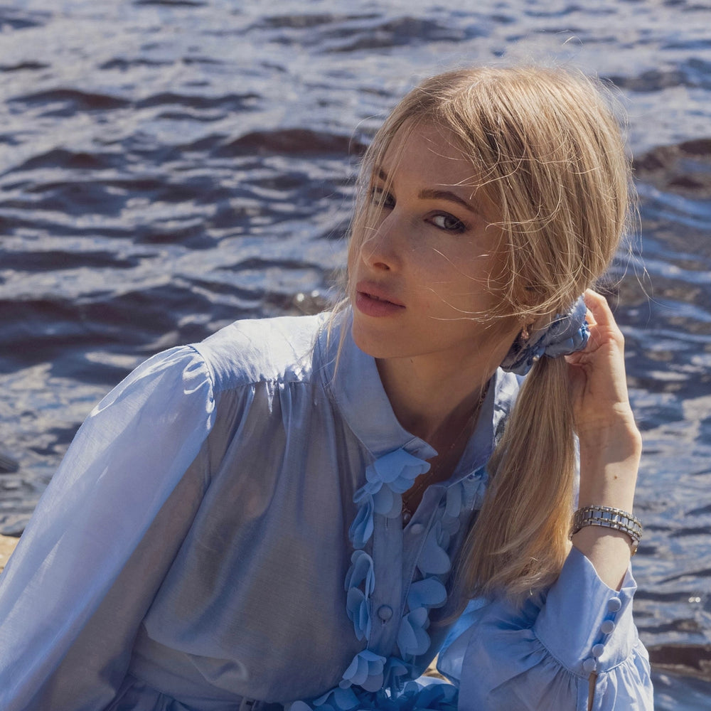 
                  
                    A blonde-haired girl in light blue dress, front view, with stylish ponytail is wearing light blue Malkiele designer silk scrunchie, embellished with pearls, name Majestic.
                  
                