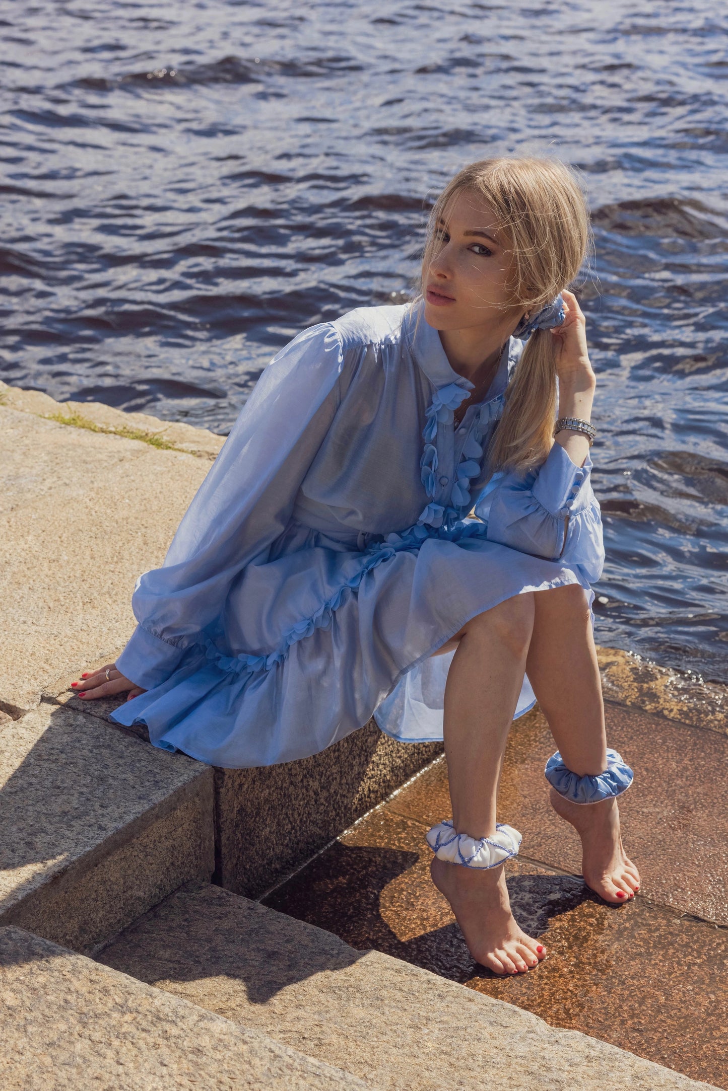 
                  
                    A blonde-haired girl in light blue dress, front view, with stylish ponytail is wearing light blue Malkiele designer silk scrunchie, embellished with pearls, name Majestic.
                  
                