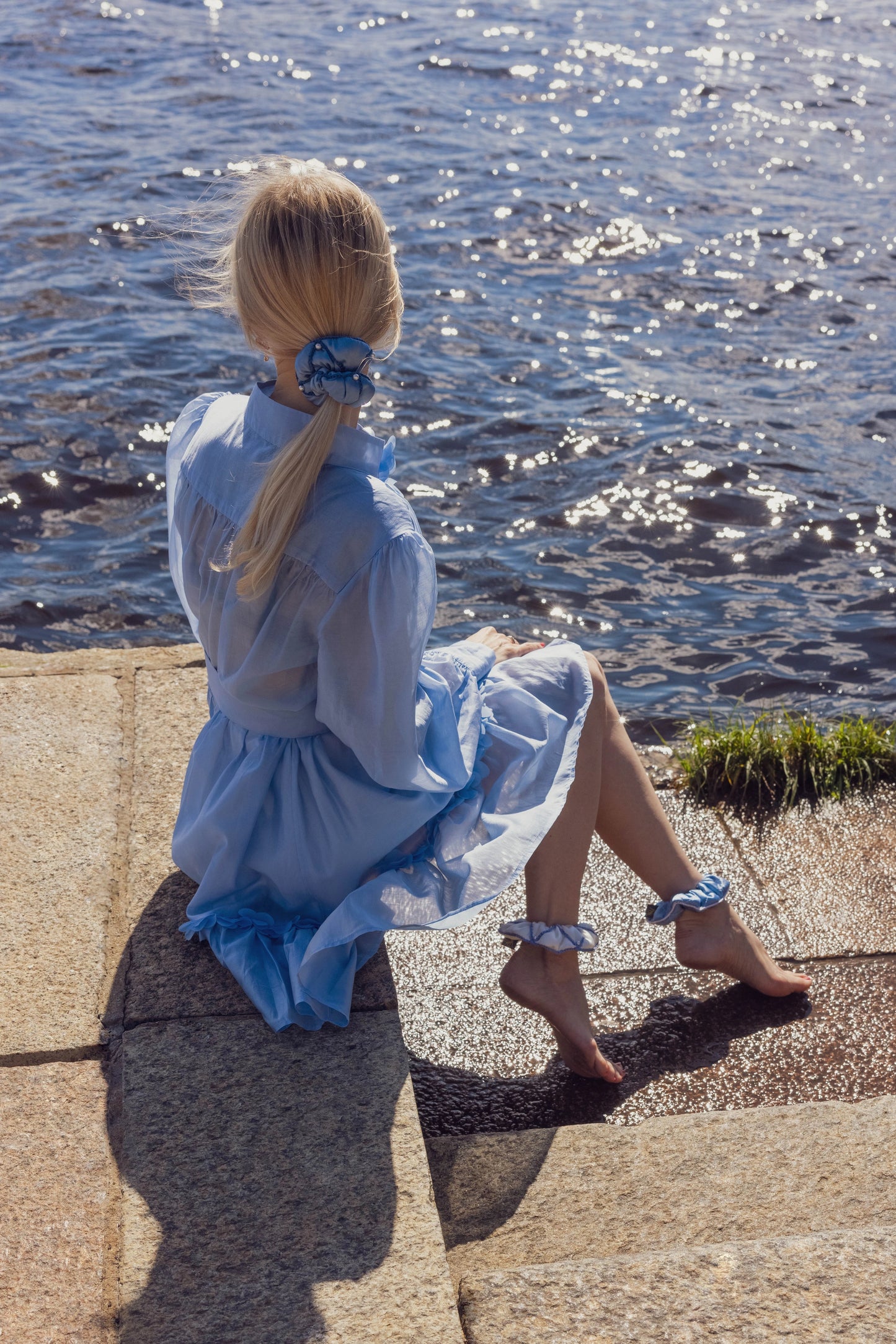 
                  
                    A blonde-haired girl in light blue dress, back view, with stylish ponytail is wearing light blue Malkiele designer silk scrunchie, embellished with pearls, name Majestic.
                  
                