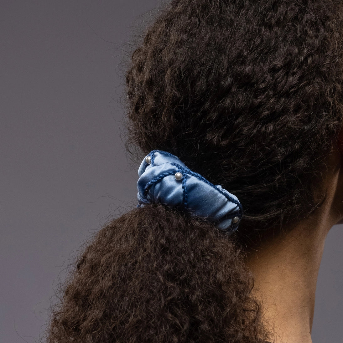 
                  
                    A dark-haired girl, back view, details, with stylish ponytail is wearing light blue Malkiele designer silk scrunchie, embellished with pearls, name Majestic.
                  
                