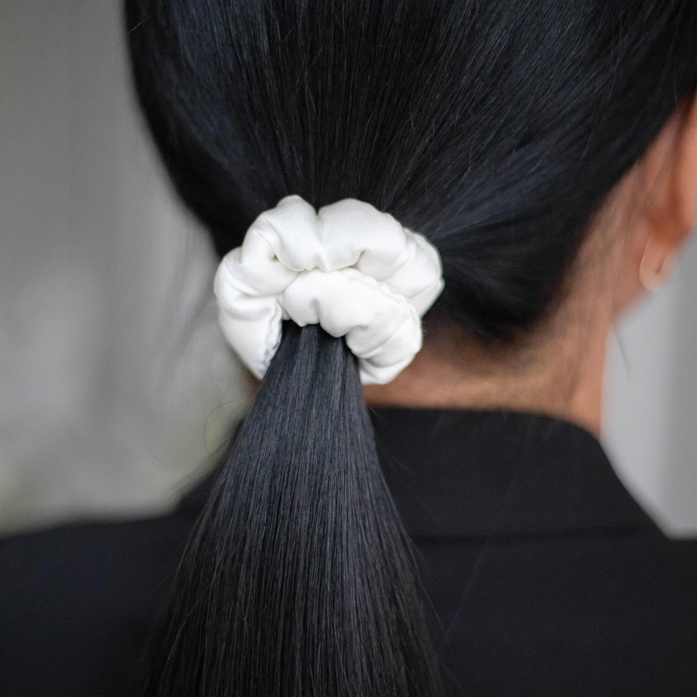 
                  
                    A dark-haired woman, view from the back, with stylish ponytail is wearing Malkiele designer white silk-covered coil scrunchie, details, name Grace.
                  
                