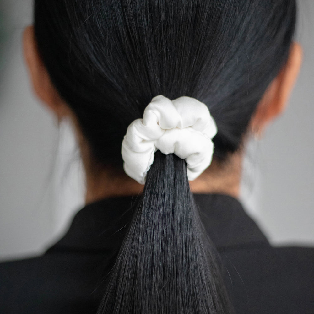 
                  
                    A dark-haired woman, view from the back, with stylish ponytail is wearing Malkiele designer white silk-covered coil scrunchie, details, name Grace.
                  
                