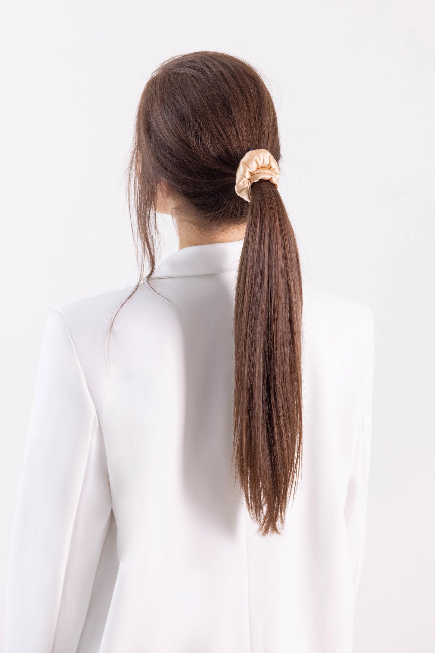 
                  
                    A dark-haired woman, view from the back, with stylish ponytail is wearing Malkiele designer beige silk-covered coil scrunchie, name Grace.
                  
                