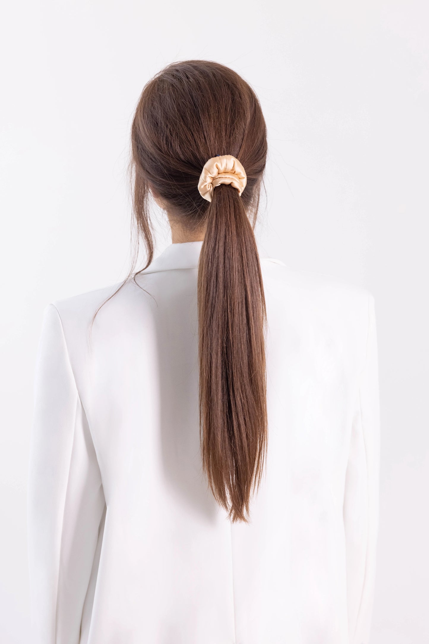 
                  
                    A dark-haired woman, view from the back, with stylish ponytail is wearing Malkiele designer beige silk-covered coil scrunchie, name Grace.
                  
                