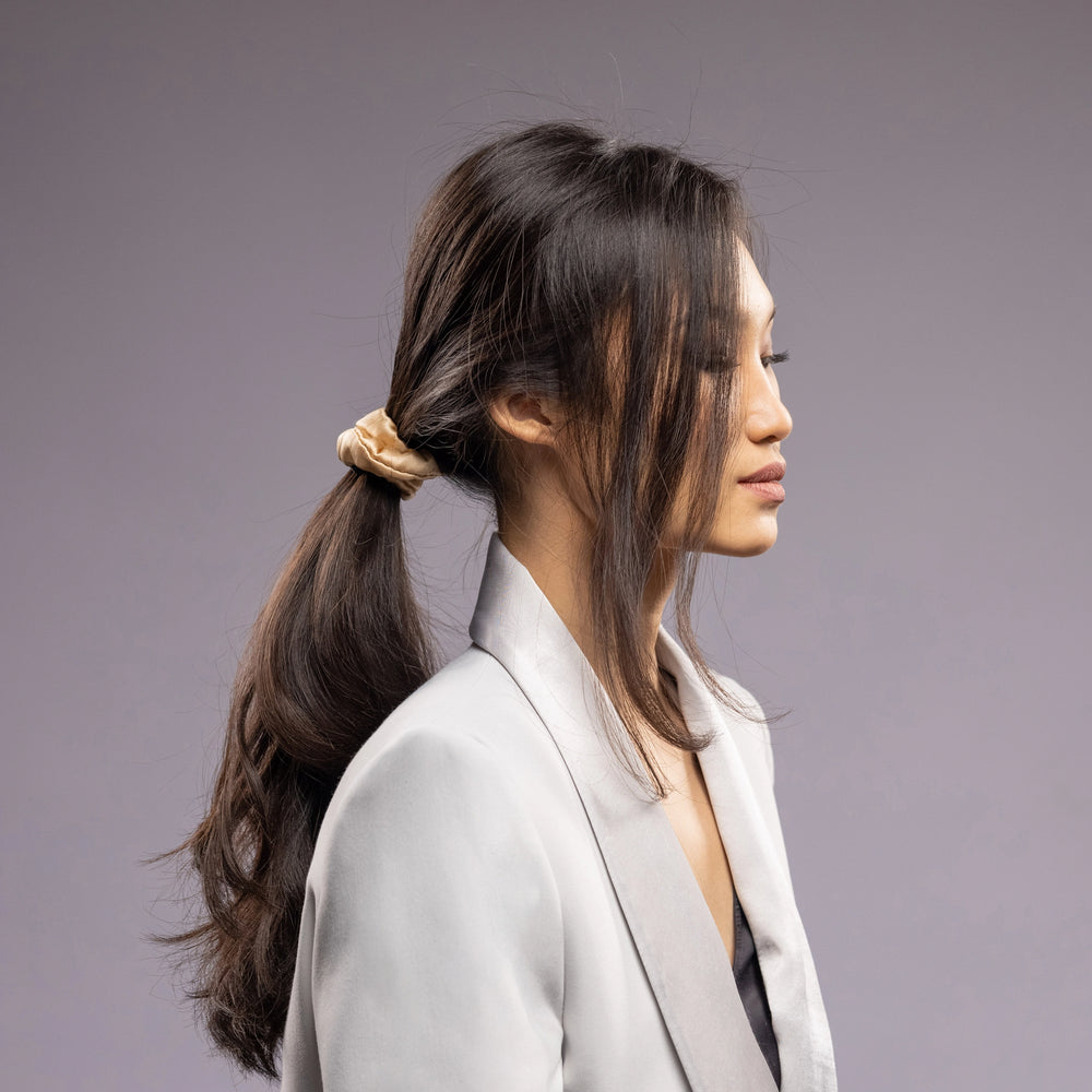 
                  
                    A black-haired woman, view from the side, with stylish ponytail is wearing Malkiele designer beige silk-covered coil scrunchie, name Grace.
                  
                