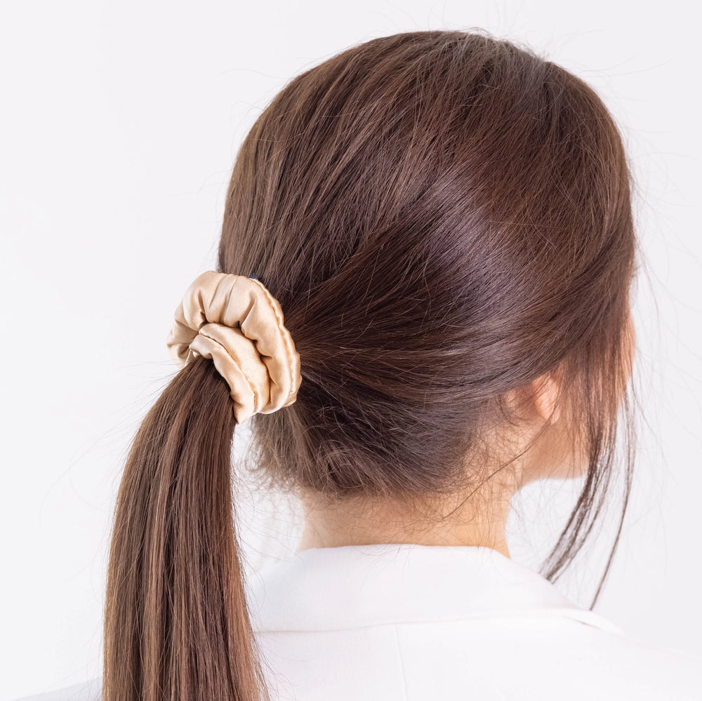 
                  
                    A dark-haired woman, view from the back, with stylish ponytail is wearing Malkiele designer beige silk-covered coil scrunchie, name Grace.
                  
                