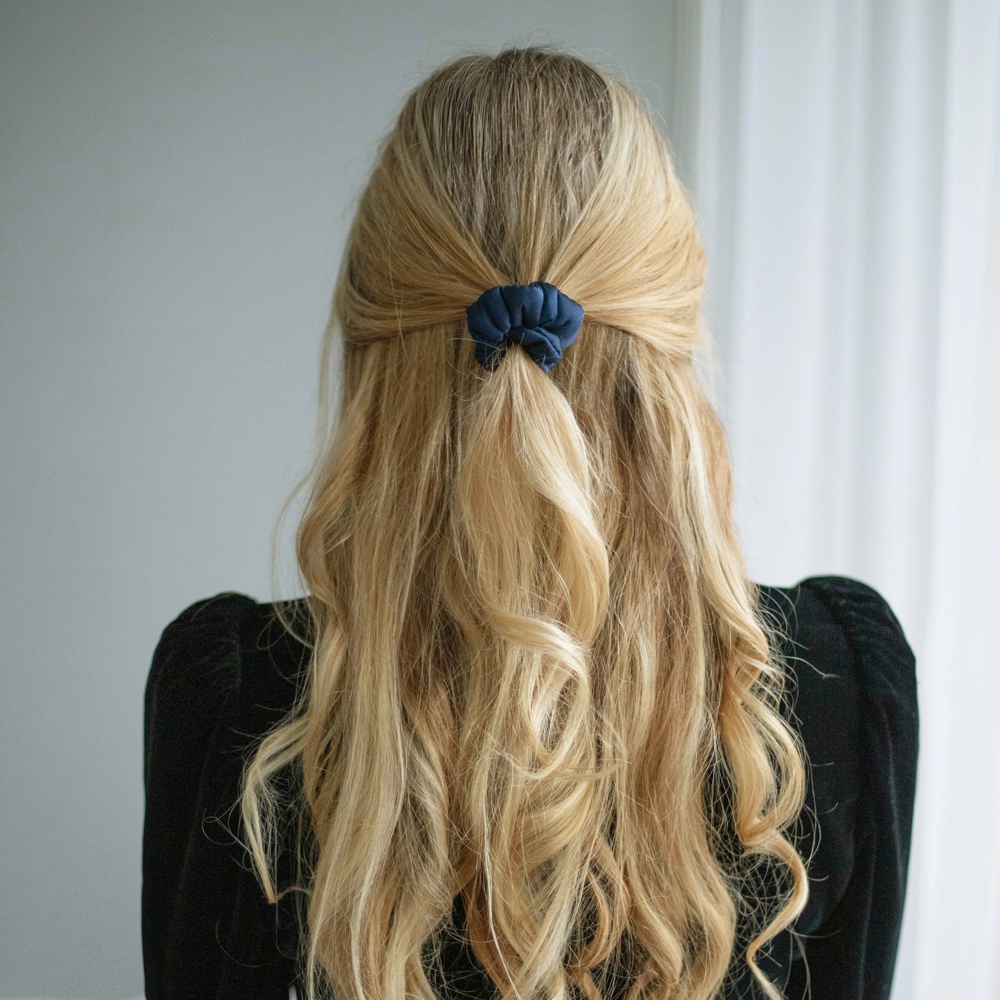 A blonde-haired woman, view from the back,  with stylish ponytail is wearing Malkiele designer navy blue silk-covered coil scrunchie, name Grace.