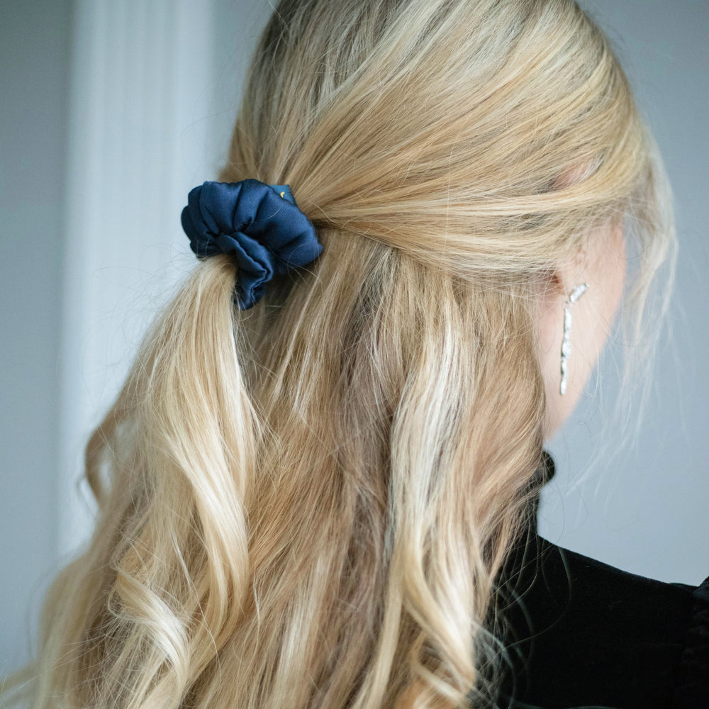 
                  
                    A blonde-haired woman, view from the back,  with stylish ponytail is wearing Malkiele designer navy blue silk-covered coil scrunchie, name Grace.
                  
                