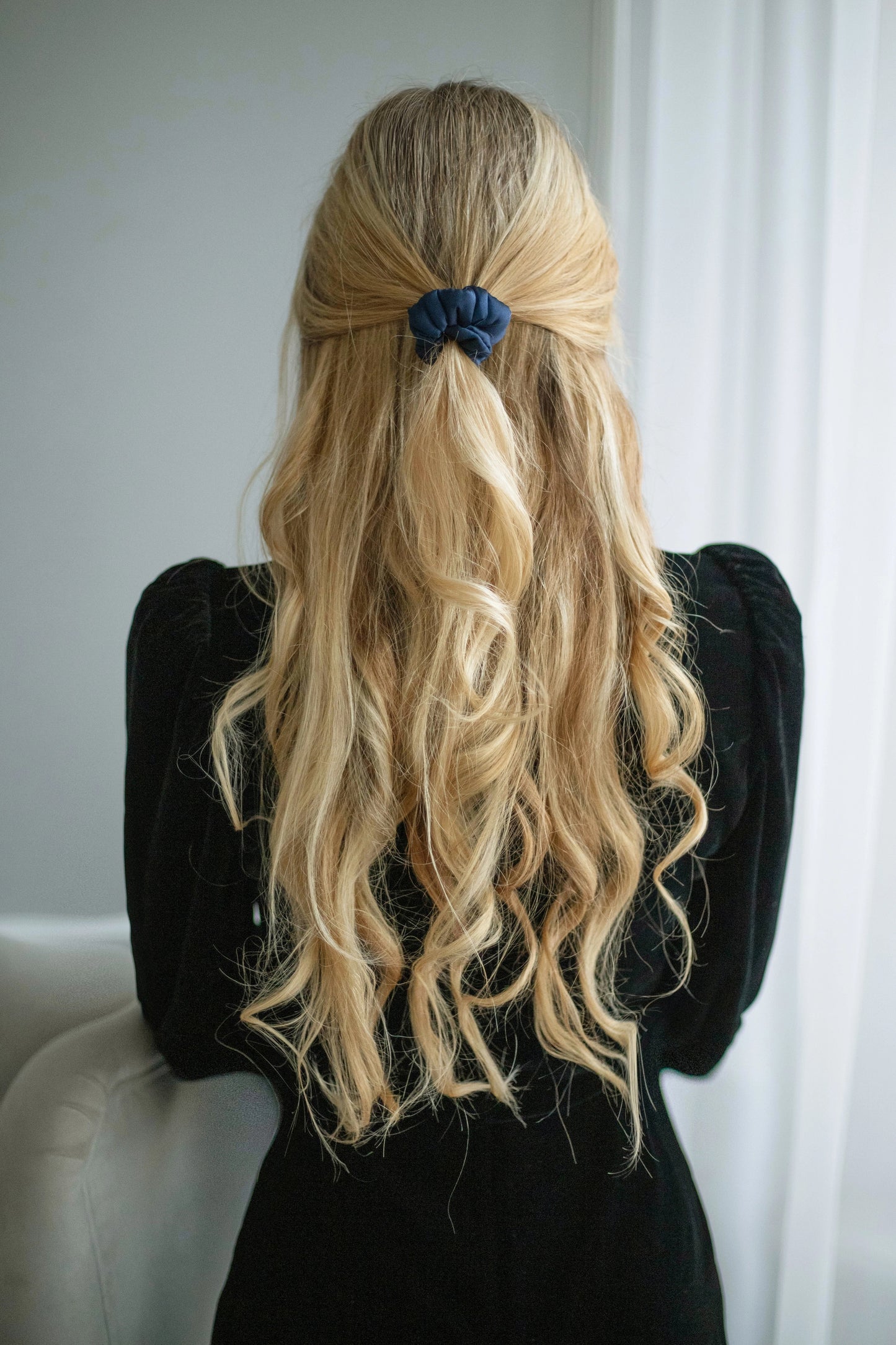 
                  
                    A blonde-haired woman, in black dress, view from the back,  with stylish ponytail is wearing Malkiele designer navy blue silk-covered coil scrunchie, name Grace.
                  
                