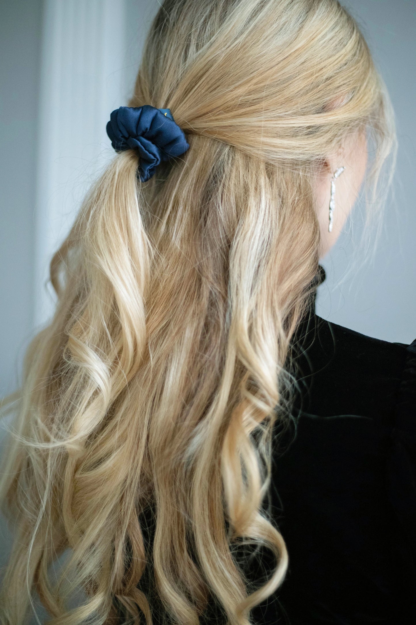 
                  
                    A blonde-haired woman, view from the back,  with stylish ponytail is wearing Malkiele designer navy blue silk-covered coil scrunchie, name Grace.
                  
                