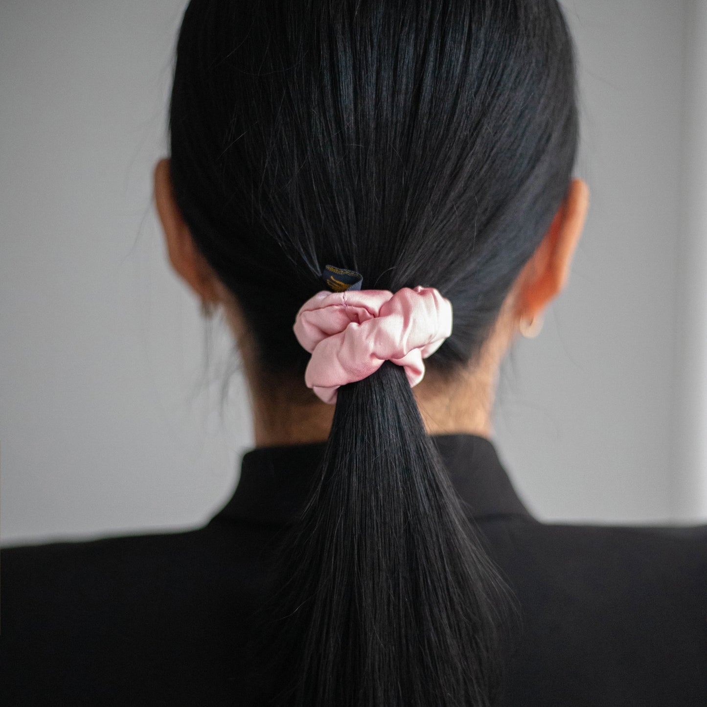 
                  
                    A black-haired woman, view from the back, with stylish ponytail is wearing Malkiele designer pink silk-covered coil scrunchie, name Grace.
                  
                