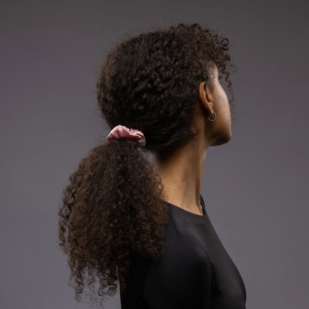 
                  
                    A dark-haired woman, view from the back, with stylish ponytail is wearing Malkiele designer pink silk-covered coil scrunchie, name Grace.
                  
                