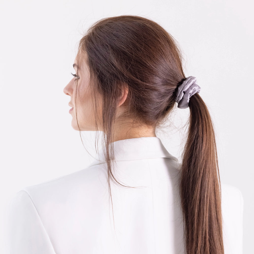 A dark-haired woman, view from the side,  with stylish ponytail is wearing Malkiele designer grey silk-covered coil scrunchie, name Grace.