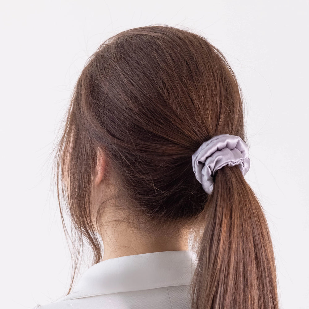 
                  
                    A dark-haired woman, back view,  with stylish ponytail is wearing Malkiele designer grey silk-covered coil scrunchie, name Grace.
                  
                