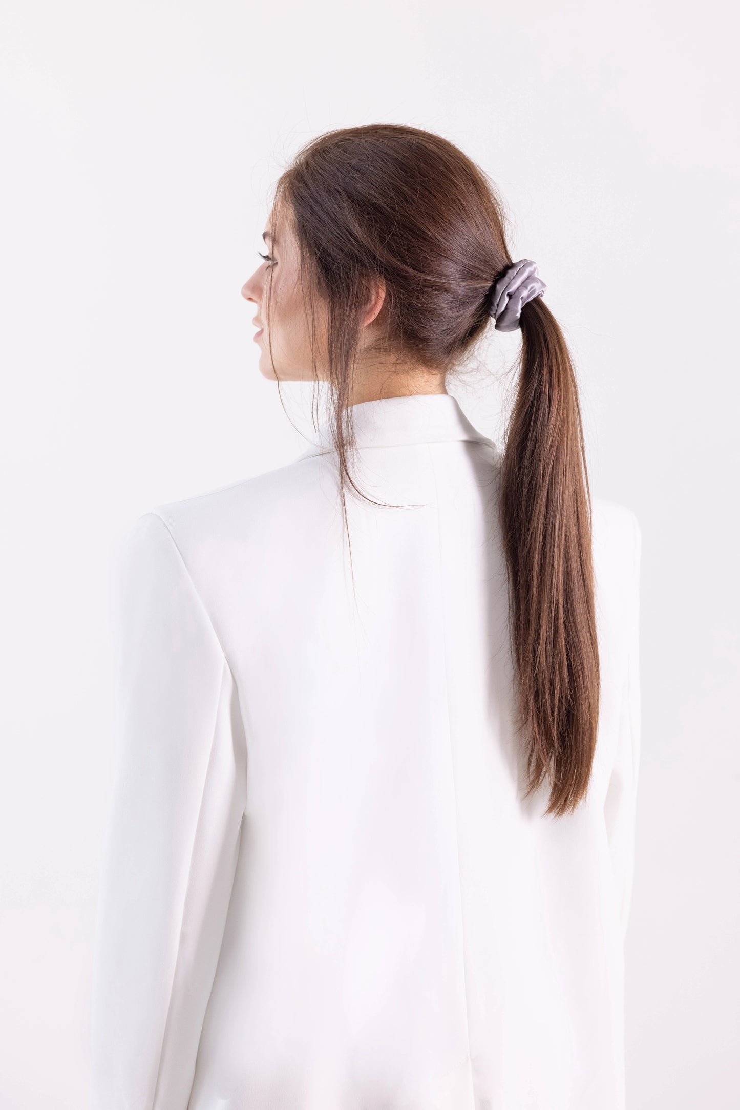 
                  
                    A dark-haired woman, view from the side,  with stylish ponytail is wearing Malkiele designer grey silk-covered coil scrunchie, name Grace.
                  
                