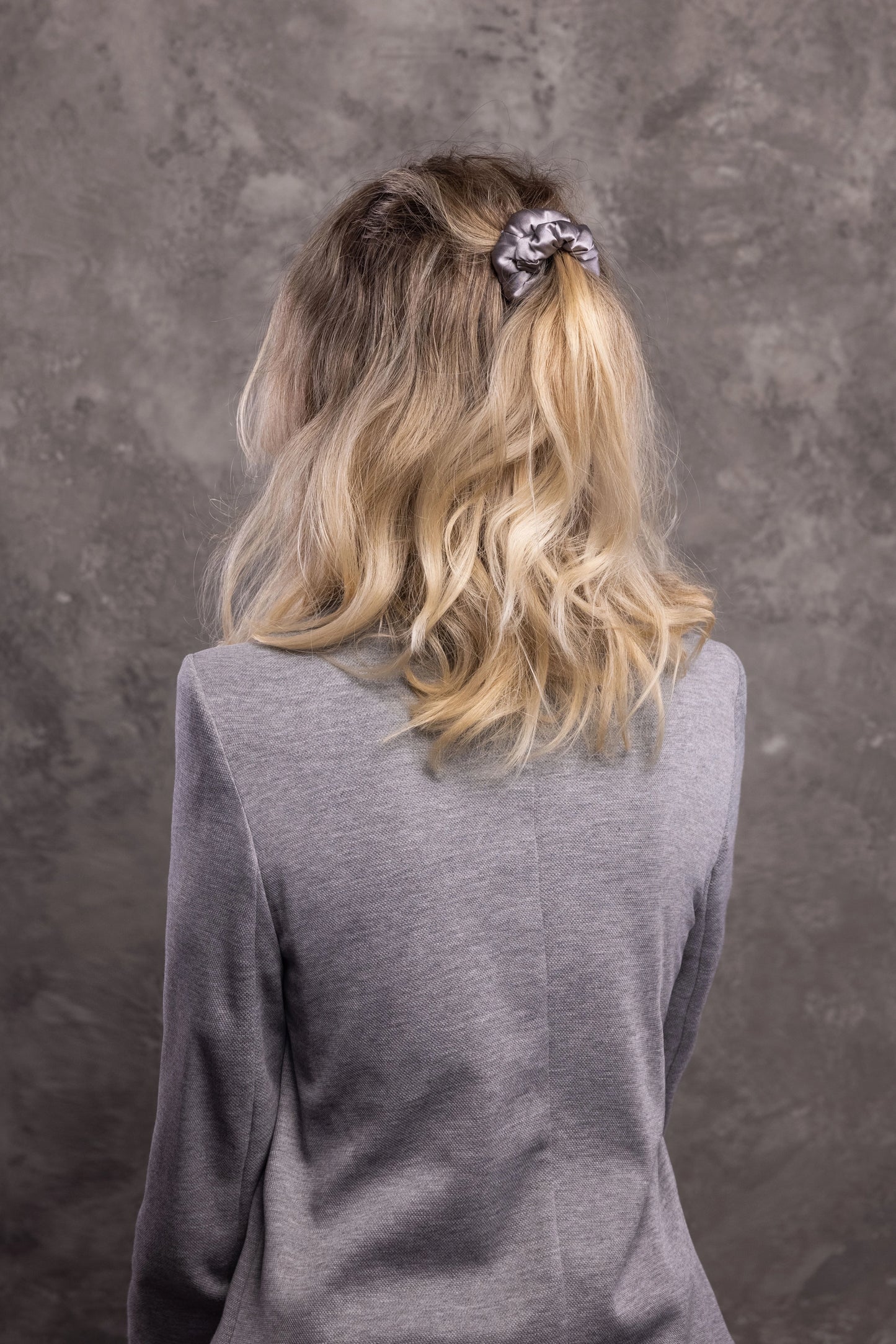 
                  
                    A blonde-haired woman, view from the back,  with stylish ponytail is wearing Malkiele designer grey silk-covered coil scrunchie, name Grace.
                  
                