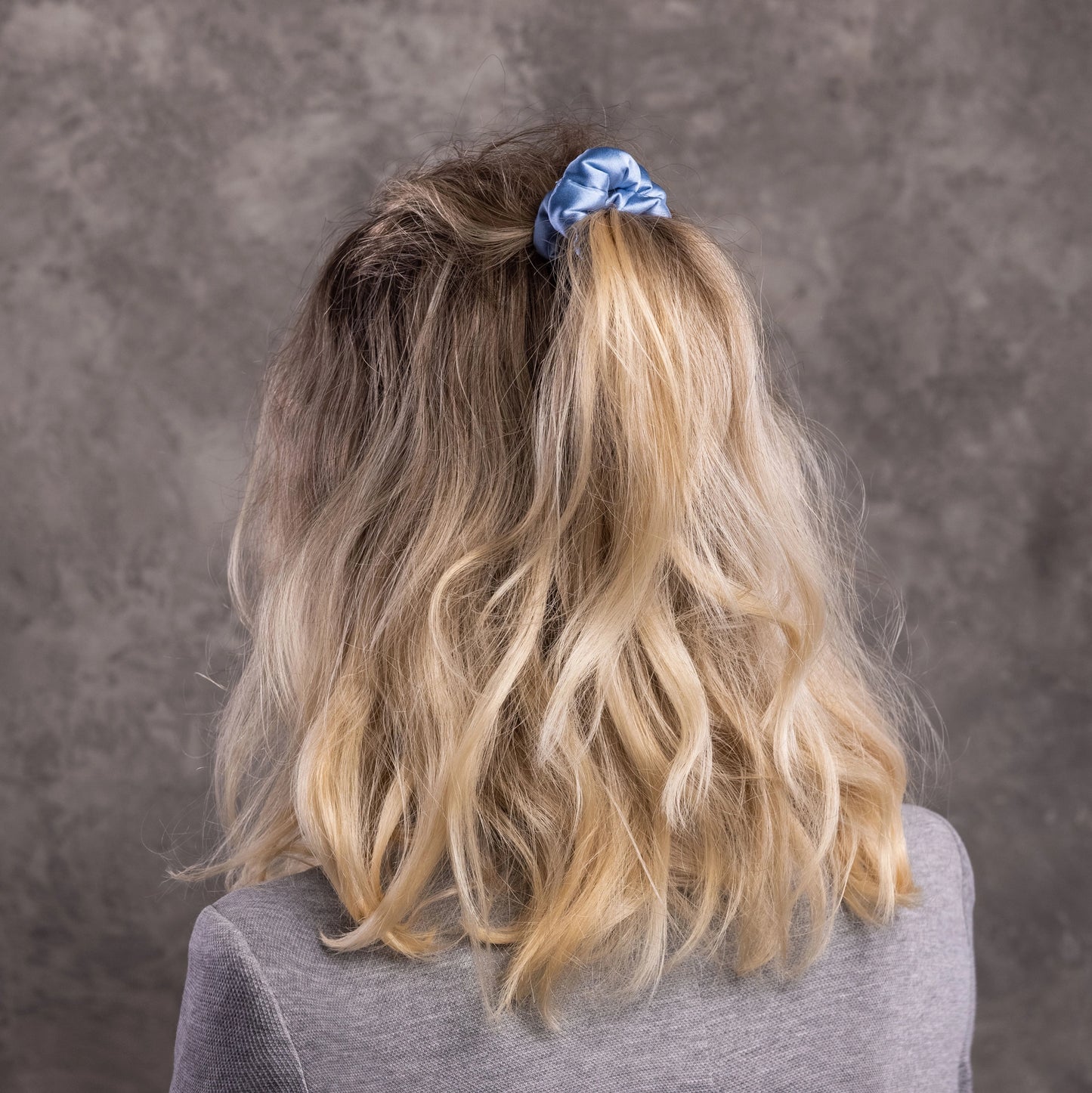 
                  
                    A blonde-haired woman, view from the back,  with stylish ponytail is wearing Malkiele designer light blue silk-covered coil scrunchie, name Grace.
                  
                