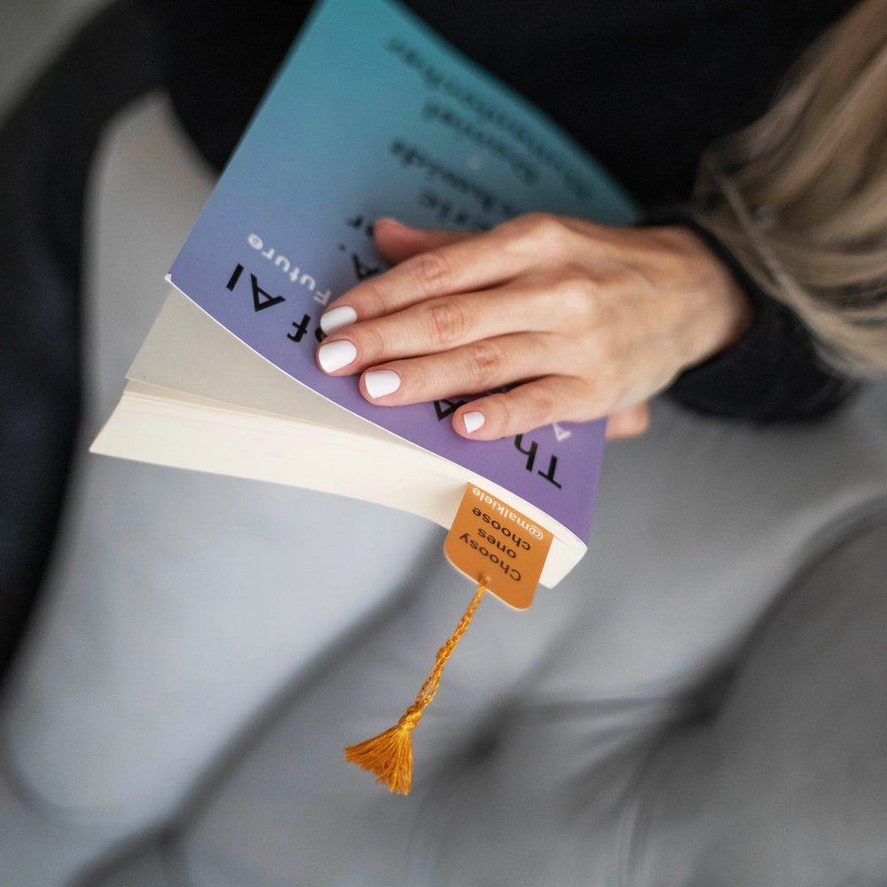 
                  
                    Woman’s hands are holding a book with Malkiele designer Bookmark with yellow silk tassel, name Witty.
                  
                