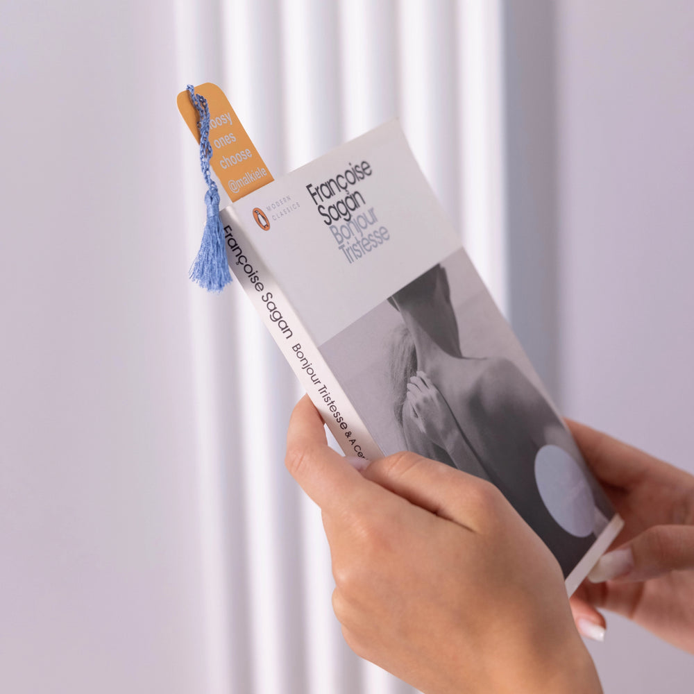 Woman hands are holding a book with yellow bookmark, embellished with a light blue silk tassel, Malkiele designer, name Witty.